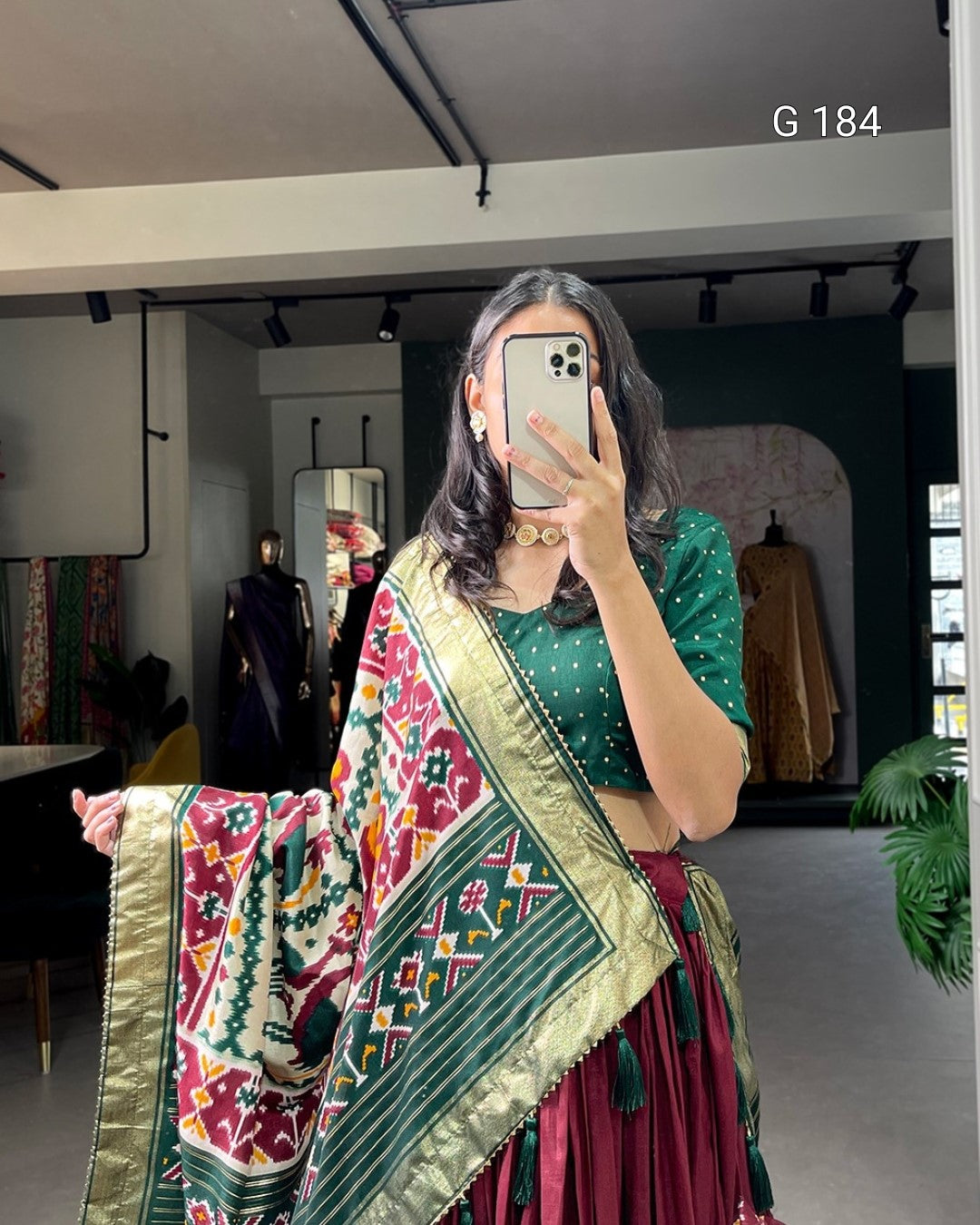 Woman taking a mirror selfie wearing a green saree with a colorful border in an indoor setting.