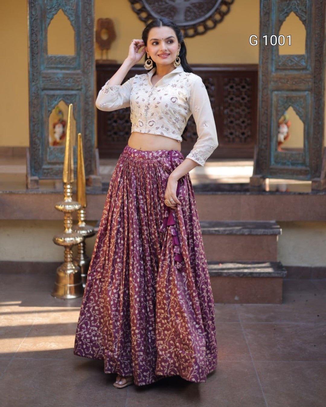 A woman modeling a traditional Indian outfit consisting of a white embroidered crop top and a long purple patterned skirt, standing in front of an ornate doorway.