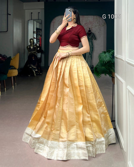 A woman takes a mirror selfie wearing a traditional Indian outfit consisting of a deep maroon short-sleeve blouse and a voluminous golden-yellow skirt with a silver border. The setting appears to be a boutique or dressing room with a mirror, plant, and furniture in the background.