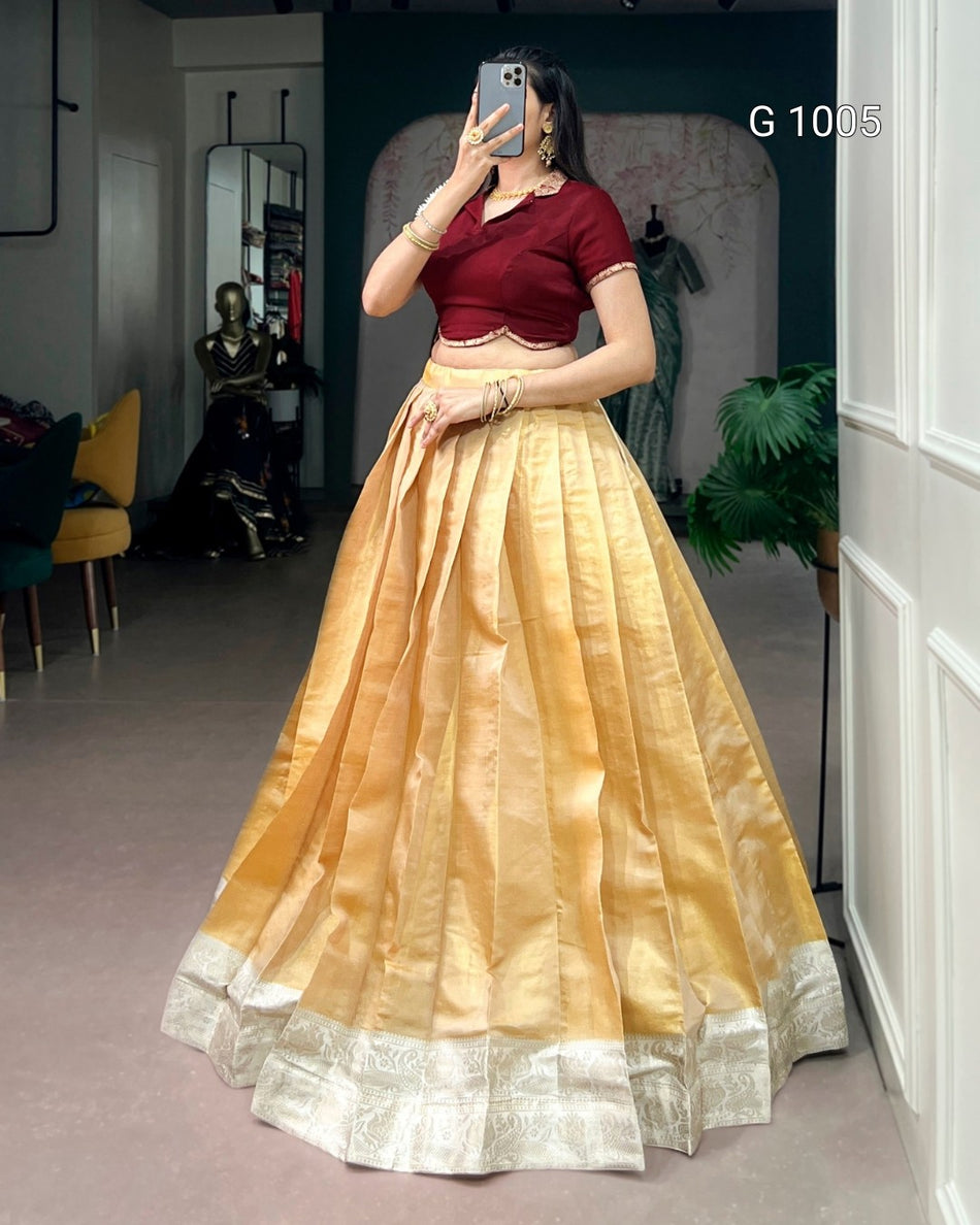 A woman takes a mirror selfie wearing a traditional Indian outfit consisting of a deep maroon short-sleeve blouse and a voluminous golden-yellow skirt with a silver border. The setting appears to be a boutique or dressing room with a mirror, plant, and furniture in the background.