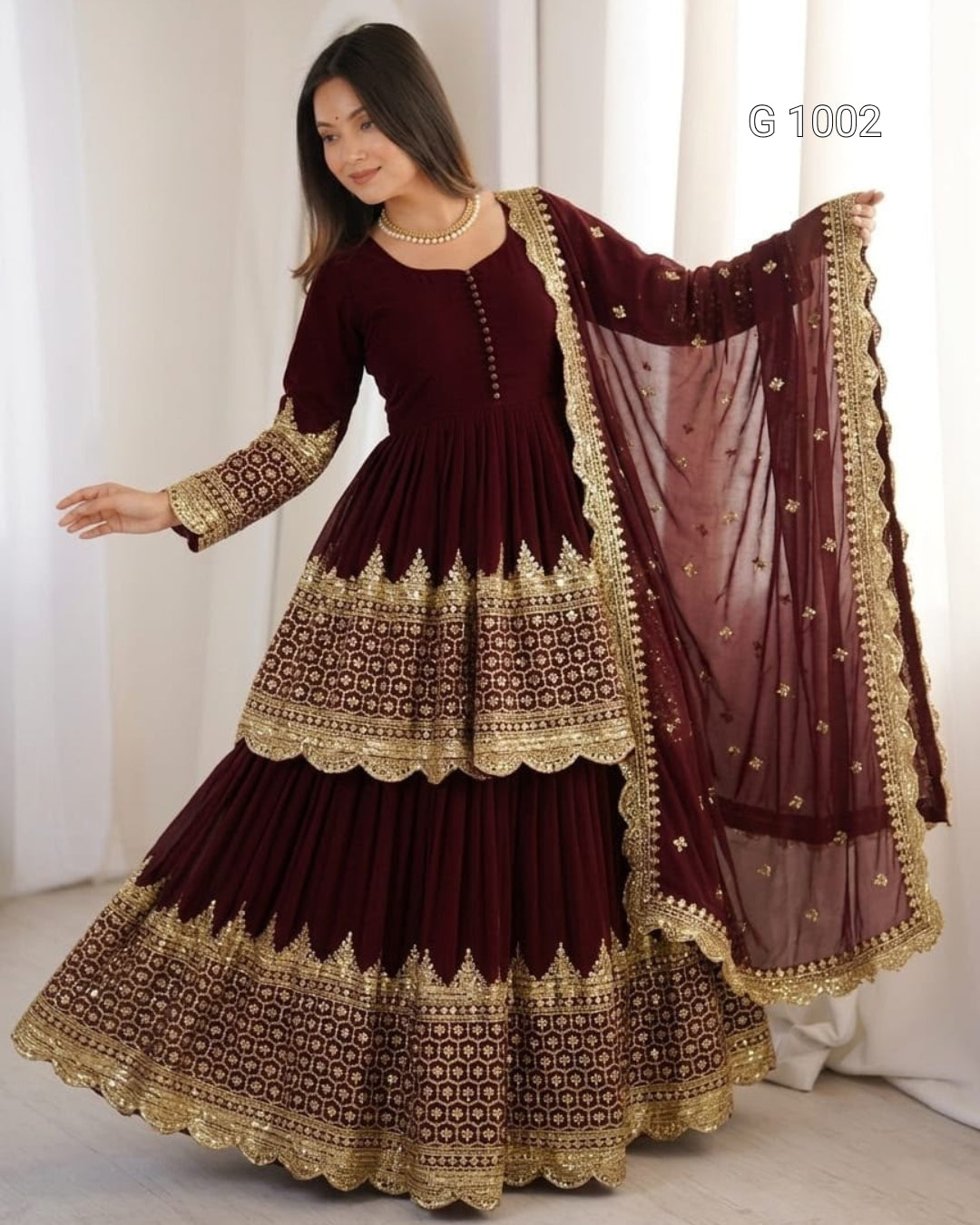 A woman modeling a traditional maroon Anarkali gown with intricate gold embroidery and a matching sheer dupatta, posed against a white curtain backdrop.