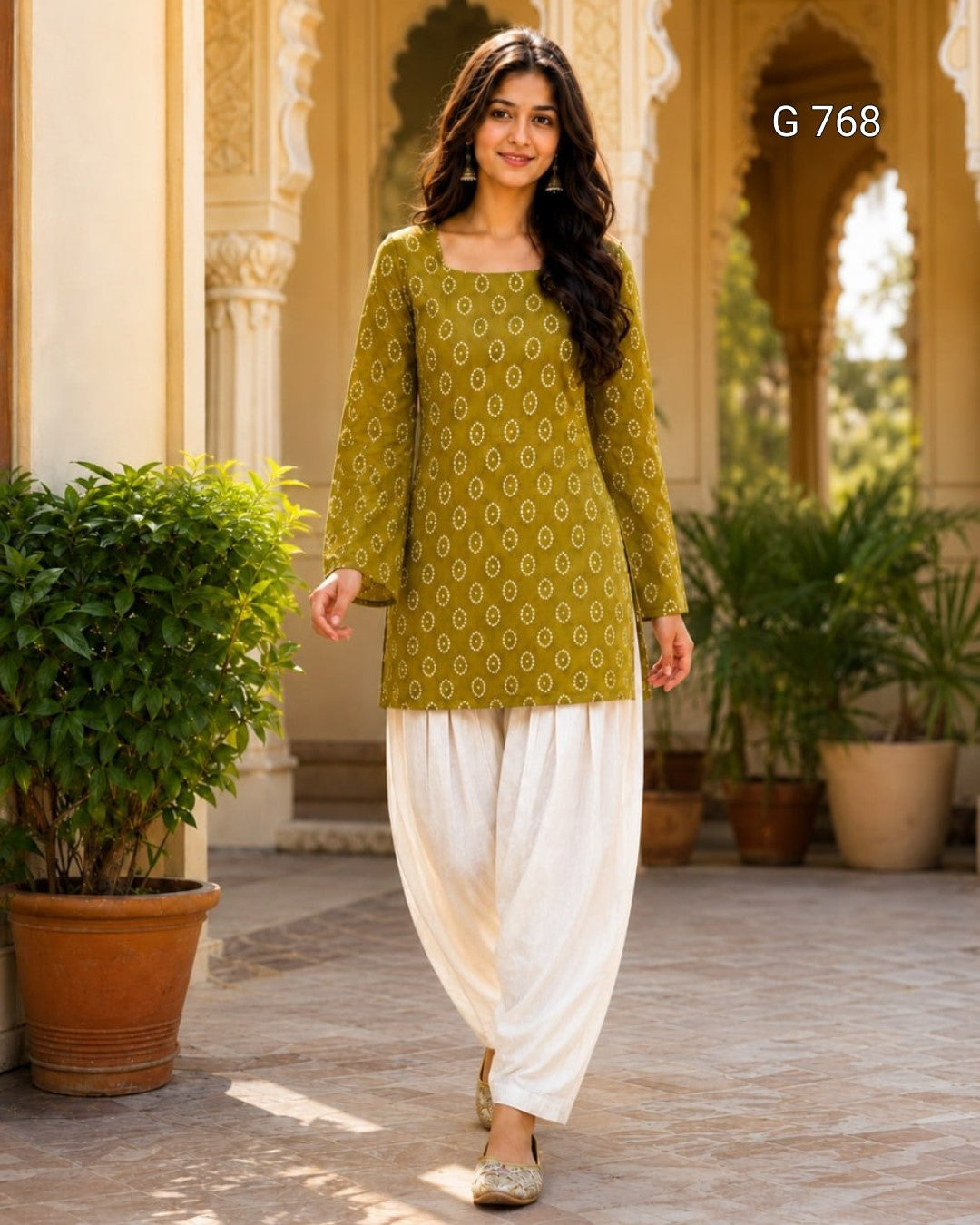 Woman wearing a green patterned top and white pants standing in front of decorative arches and plants.