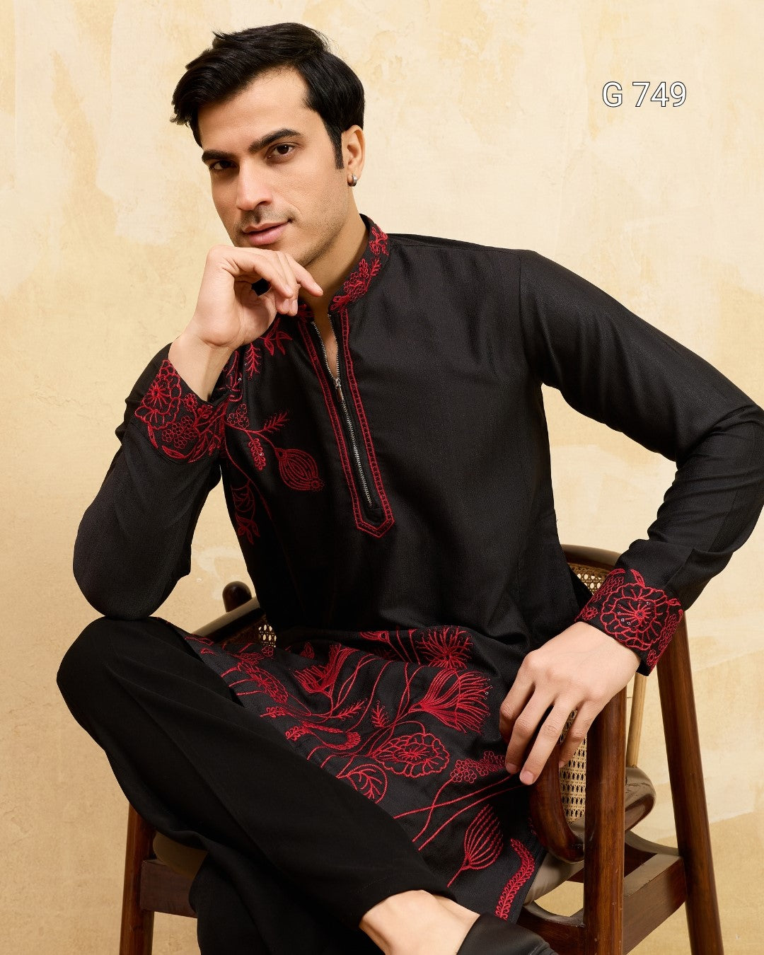 Man wearing a black kurta with red embroidery sitting on a chair against a beige background
