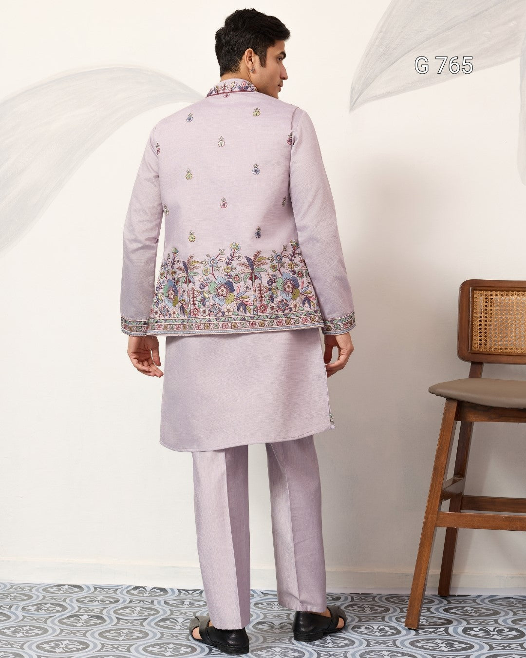Person wearing a light purple traditional outfit with intricate designs, standing against a white wall.