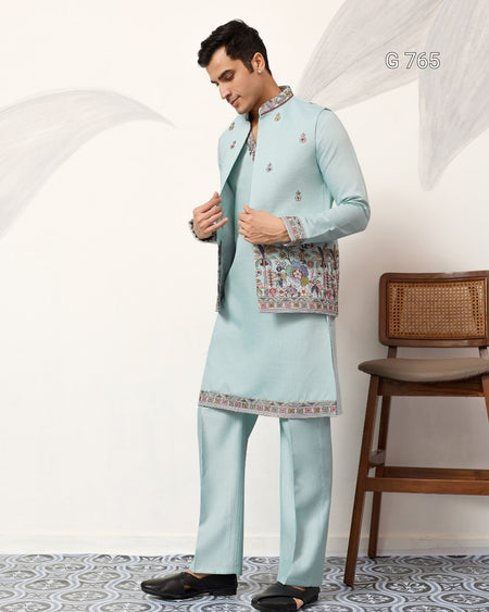 Man wearing a light blue traditional outfit with a patterned jacket in a minimalistic room.