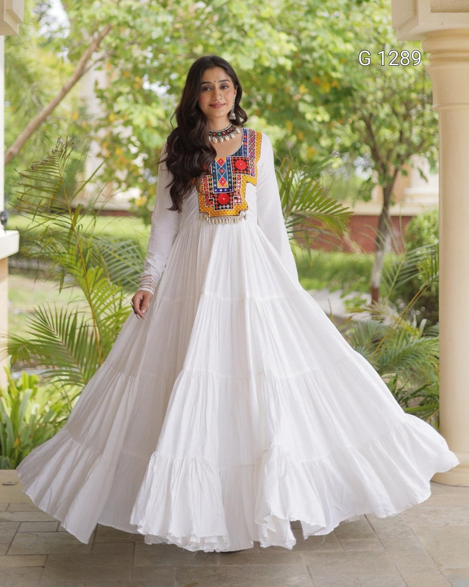 A woman wearing a white tiered maxi dress with colorful geometric embroidery around the neckline, standing outdoors in front of a garden and architectural background.