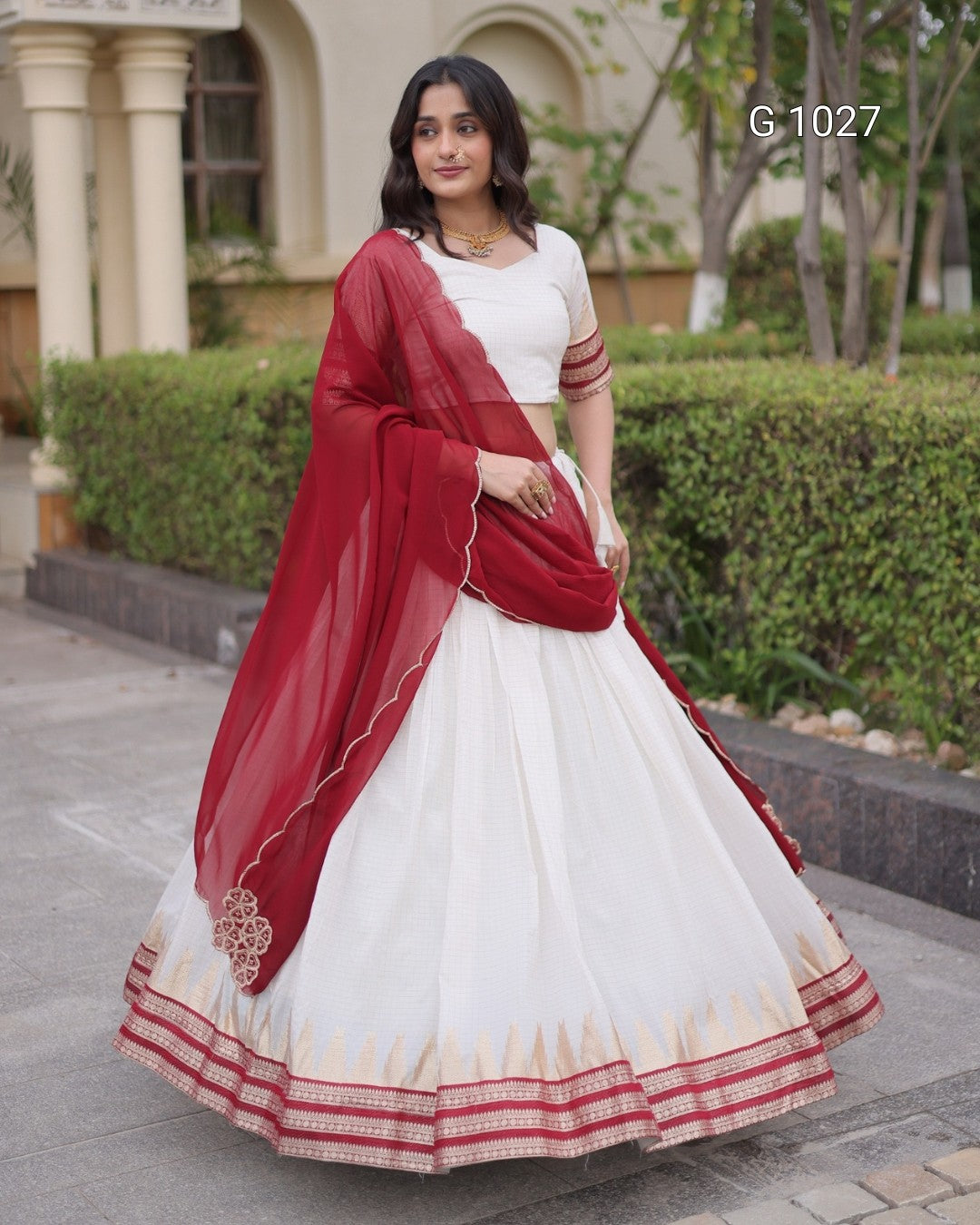 A woman modeling a traditional Indian outfit consisting of a white flared skirt (lehenga), a white sleeveless top, and a deep red dupatta, standing outdoors in front of a building with green hedges. The text 'G 1027' is visible in the top right corner.