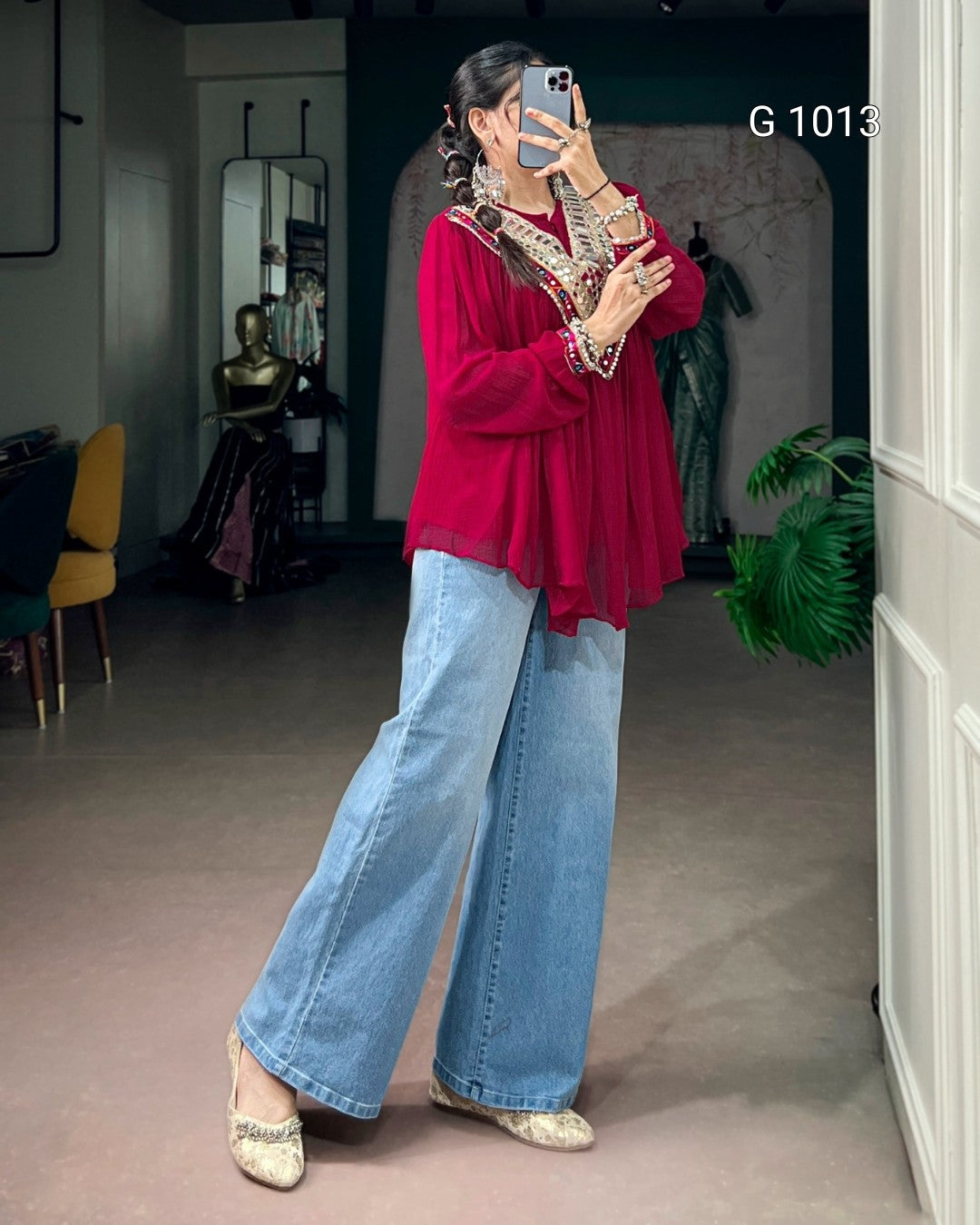 A woman takes a mirror selfie in a boutique, wearing a vibrant magenta long-sleeve blouse with gold embroidery, light-wash wide-leg denim jeans, and beige pointed flats. The boutique interior features a dark teal wall, a mannequin, and a potted green plant.