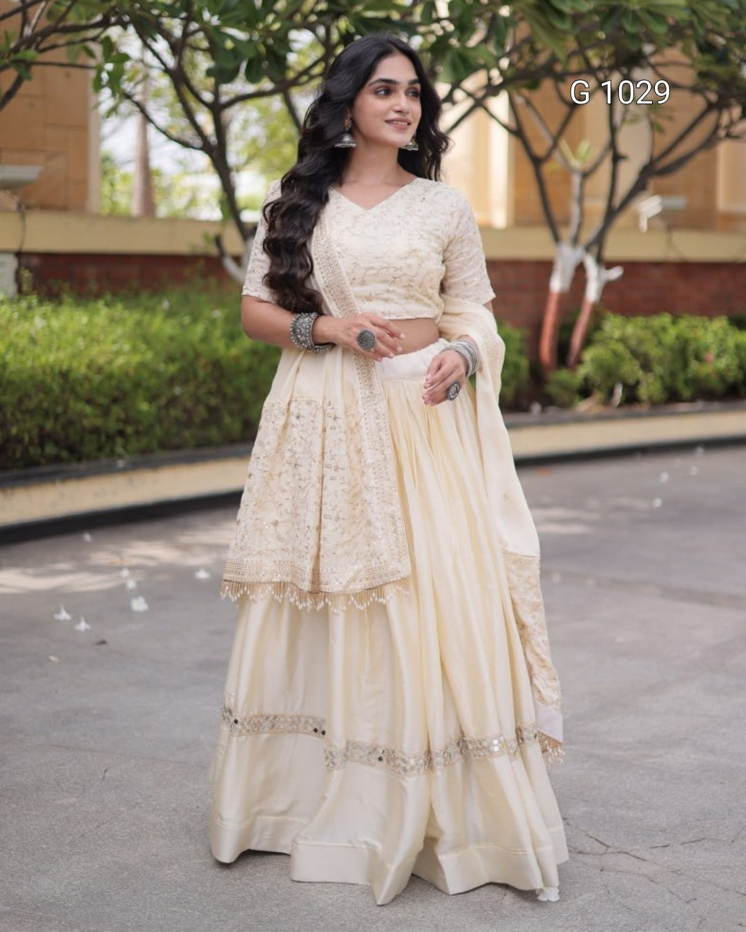 A woman wearing a cream-colored traditional Indian ethnic outfit with a heavily embroidered blouse, flared skirt, and matching dupatta, standing outdoors on a paved walkway with trees and a building in the background.