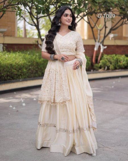 A woman wearing a cream-colored traditional Indian ethnic outfit with a heavily embroidered blouse, flared skirt, and matching dupatta, standing outdoors on a paved walkway with trees and a building in the background.