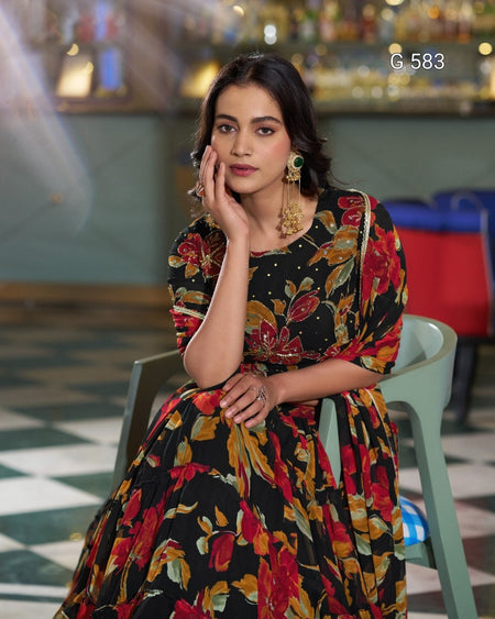 A woman wearing a black floral print dress with red, orange, and yellow flowers, sitting on a light green plastic chair in an indoor setting with a blurred background.