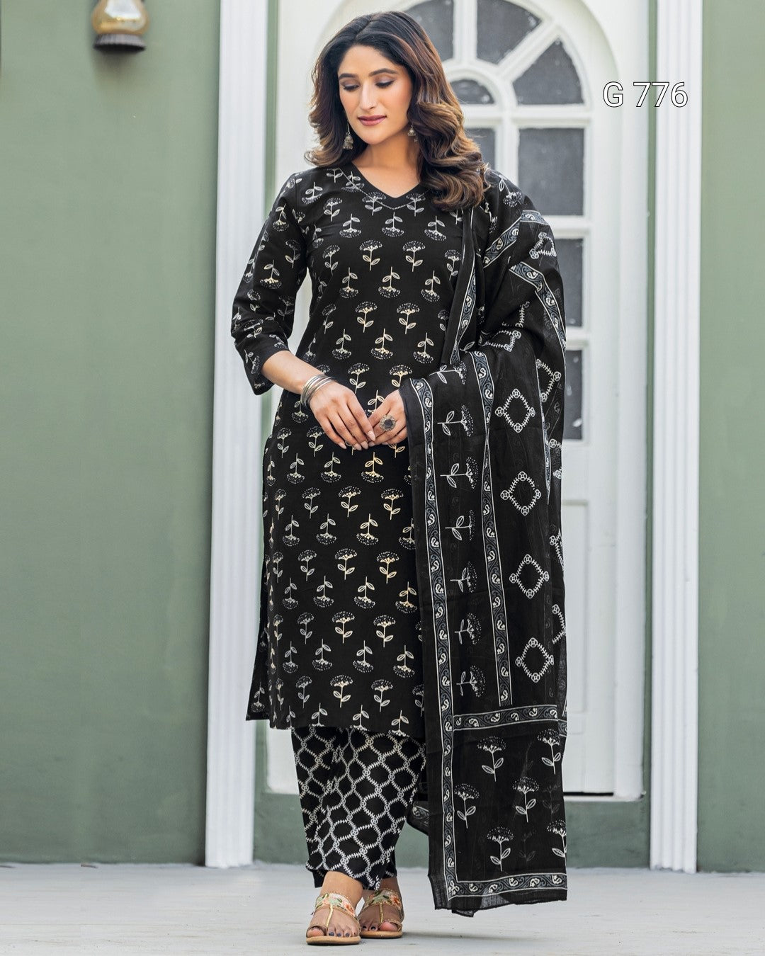 Woman wearing a black patterned outfit with a matching dupatta in front of a white door.