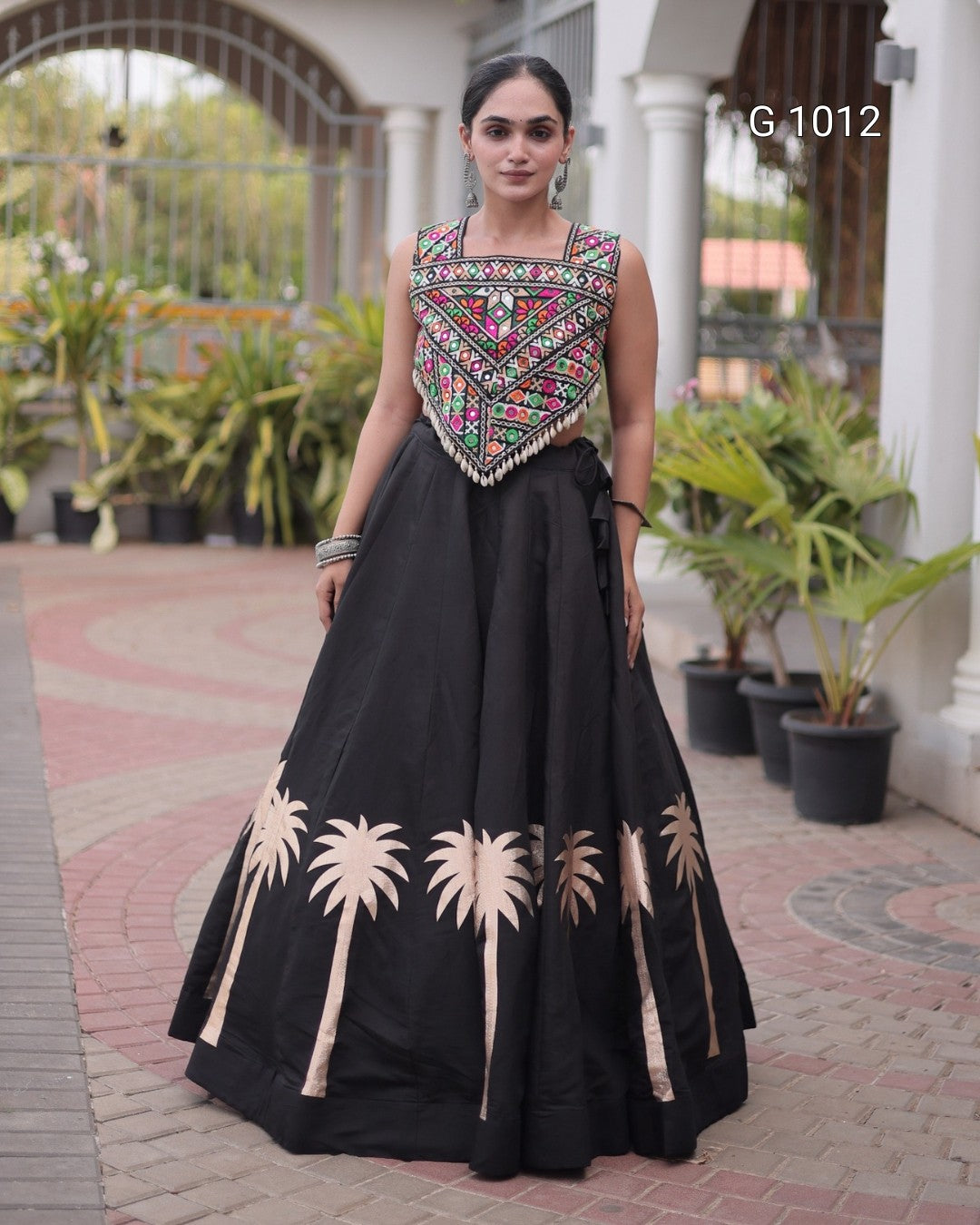 A woman modeling a black traditional Anarkali gown with a heavily embroidered multicolor bodice and beige palm tree motifs near the hem, standing outdoors on a paved walkway.
