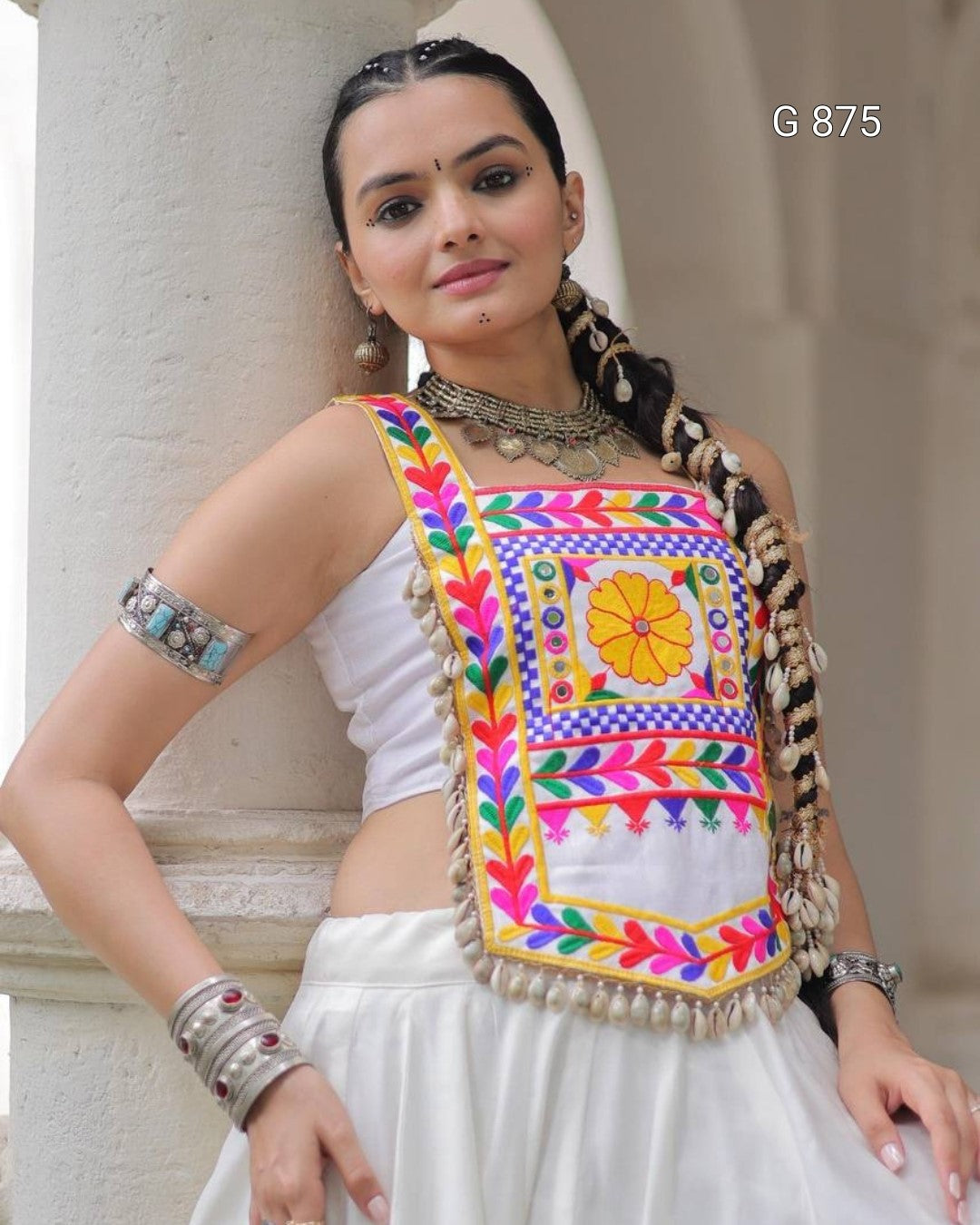 Woman wearing a colorful embroidered blouse with a white saree, standing against a neutral background.