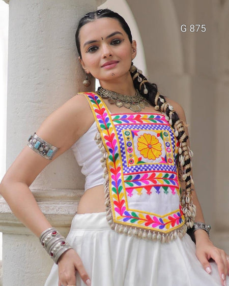 Woman wearing a colorful embroidered blouse with a white saree, standing against a neutral background.