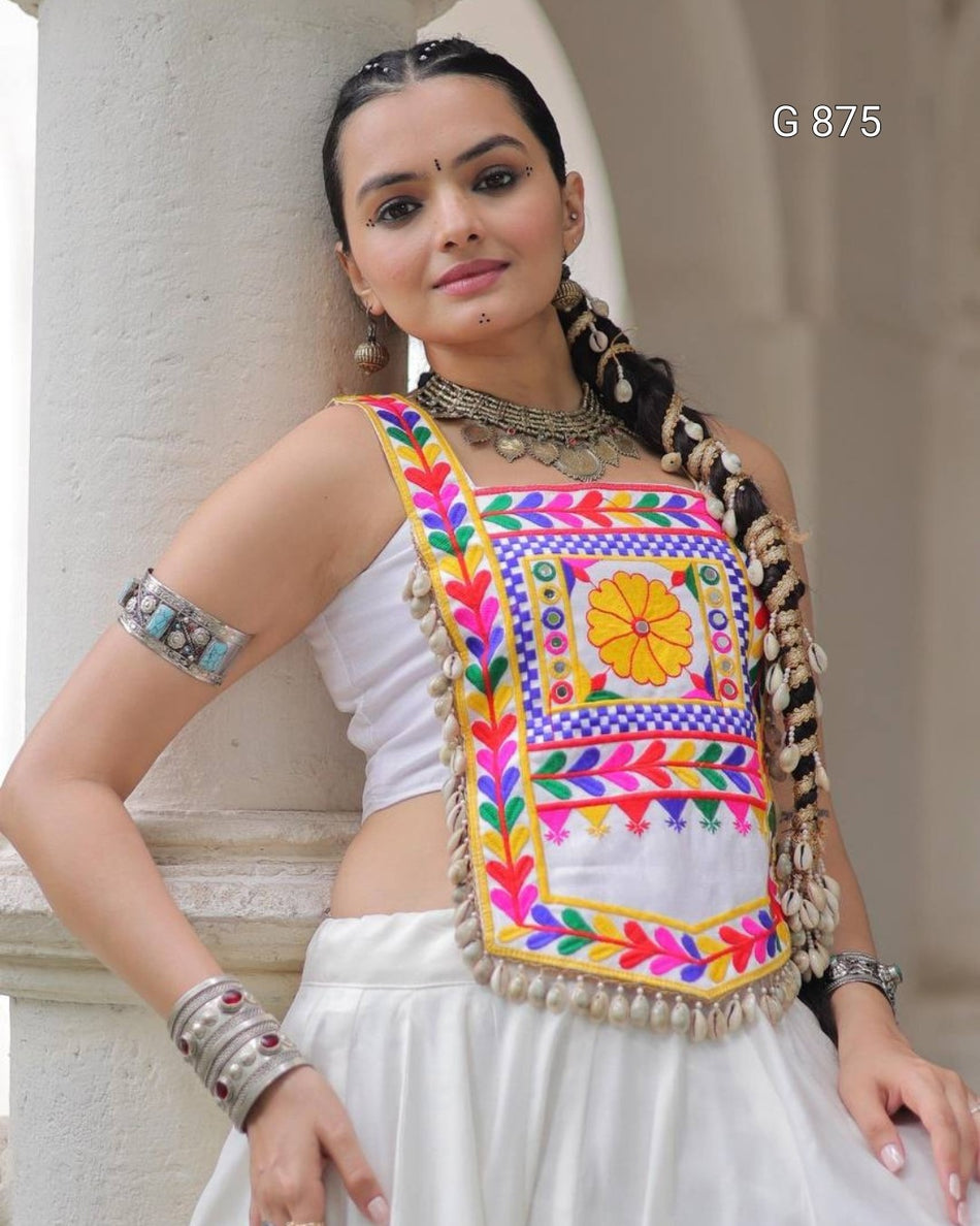 Woman wearing a colorful embroidered blouse with a white saree, standing against a neutral background.