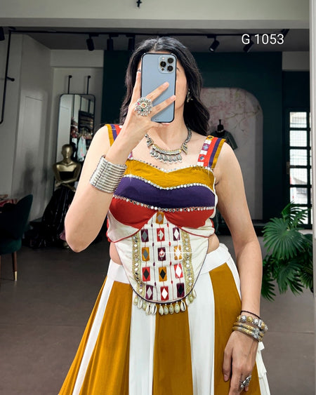 A woman takes a mirror selfie wearing a highly embellished, multi-colored traditional-style dress with a mustard yellow, white, red, and purple bodice, silver jewelry, and a decorative embroidered waist panel. The background is a boutique interior with dark green walls and mirrors.