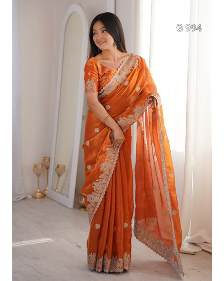 A woman modeling an orange saree with intricate silver floral embroidery and a matching sheer dupatta, standing in a bright, modern room with a large mirror and white curtains.