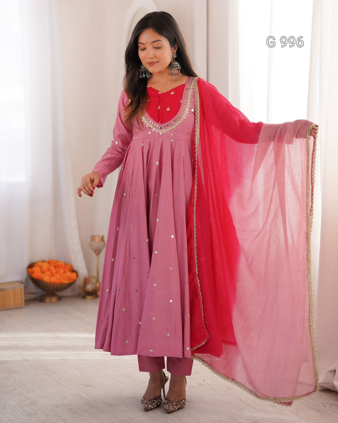 A woman models a traditional Indian outfit consisting of a dusty pink long kurta with silver mirror work, a bright pink inner bodice, matching trousers, and a sheer pink dupatta with gold border detailing, set against a soft white curtain background.