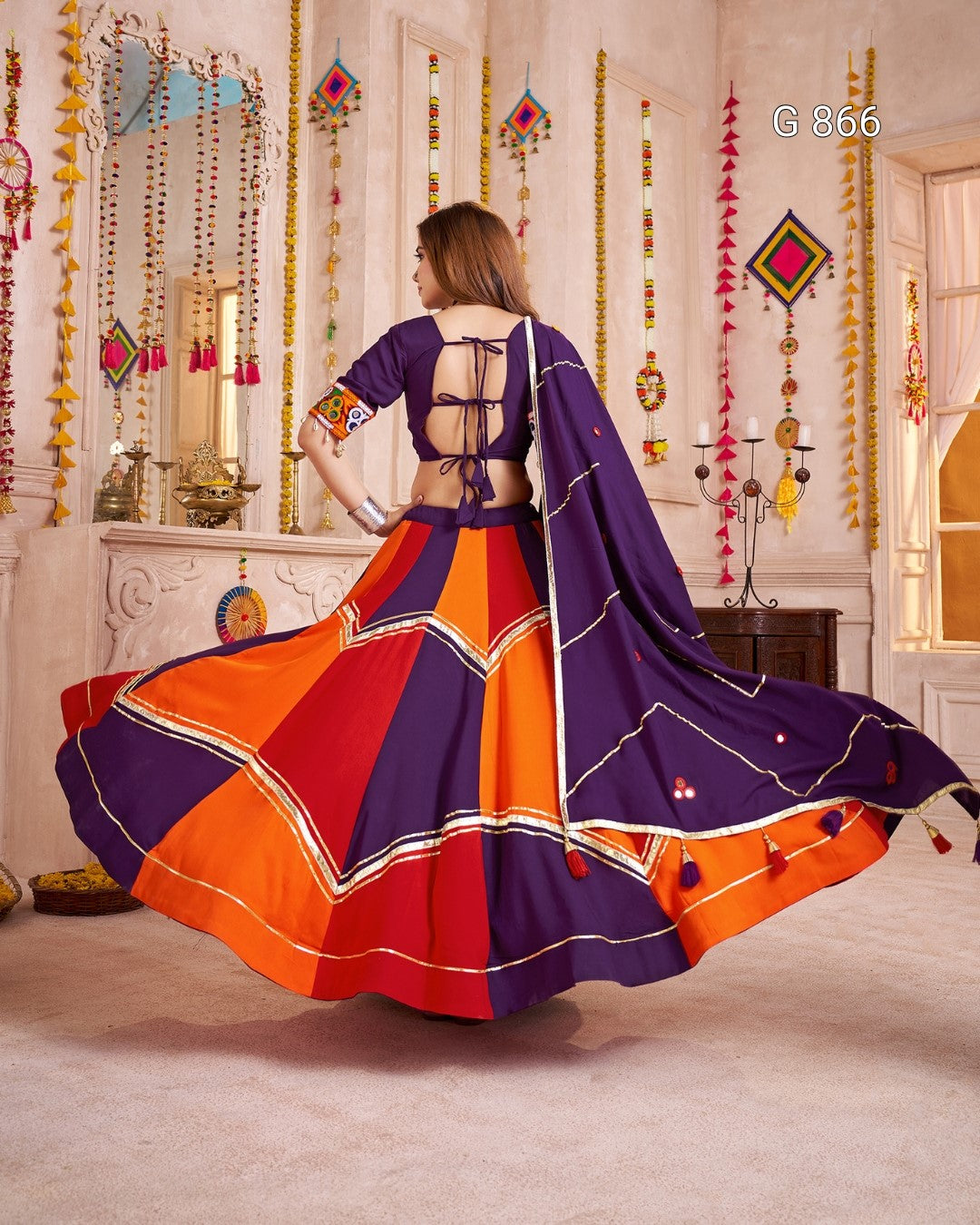 Woman in a colorful traditional outfit with a vibrant purple and orange design, standing in a decorated room.