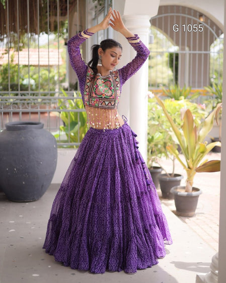 A woman modeling a purple embroidered traditional outfit with a long pleated skirt and embroidered bodice, standing in a covered outdoor walkway with potted plants.