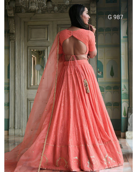 A woman wearing a peach-pink traditional Indian lehenga choli dress with a high open back, shown from behind in an ornate indoor setting. The dress features a flared skirt, short sleeves, and a sheer dupatta with gold embroidery.