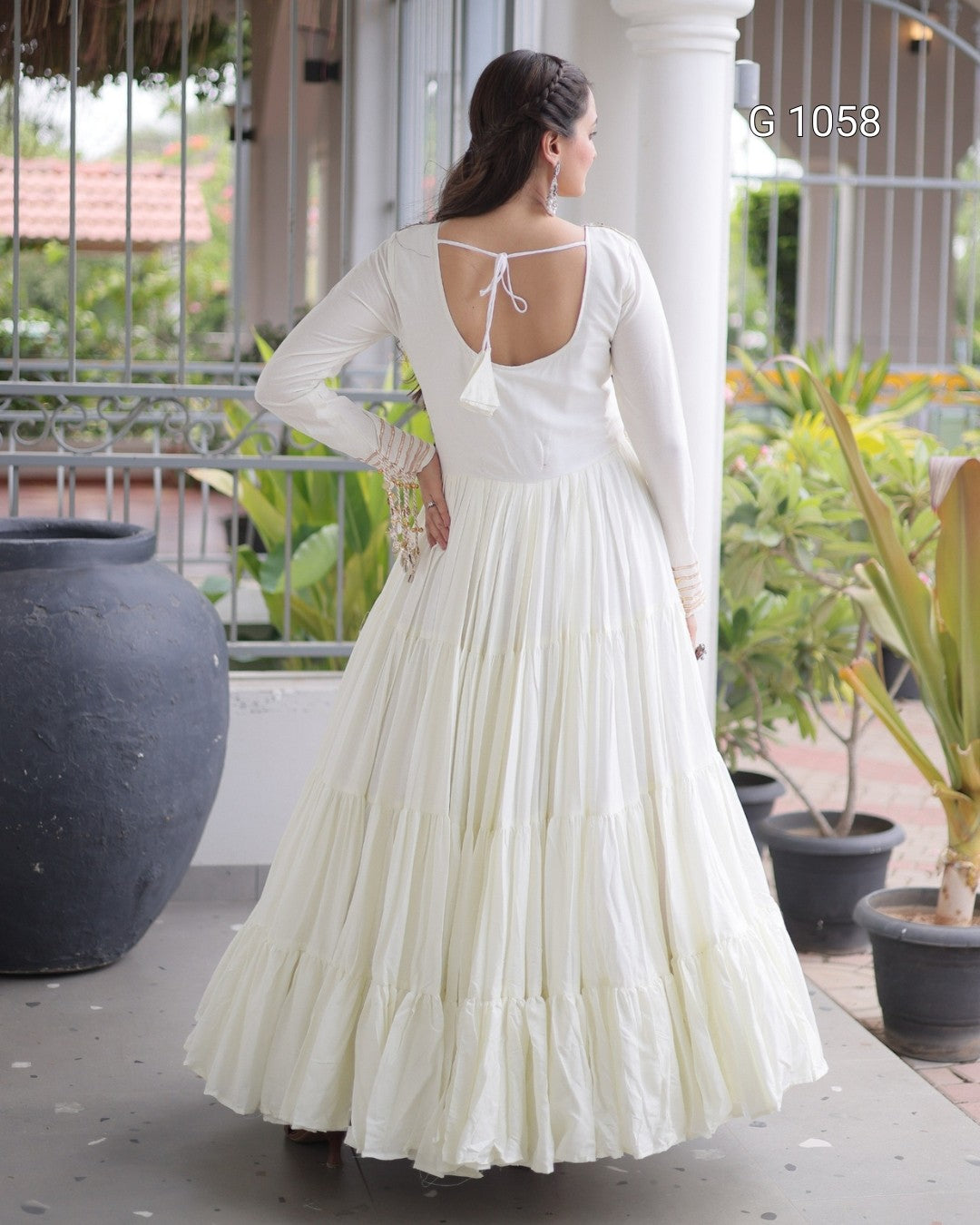 A woman wearing a long-sleeved white tiered maxi dress, shown from the back on an outdoor porch with potted plants and columns.