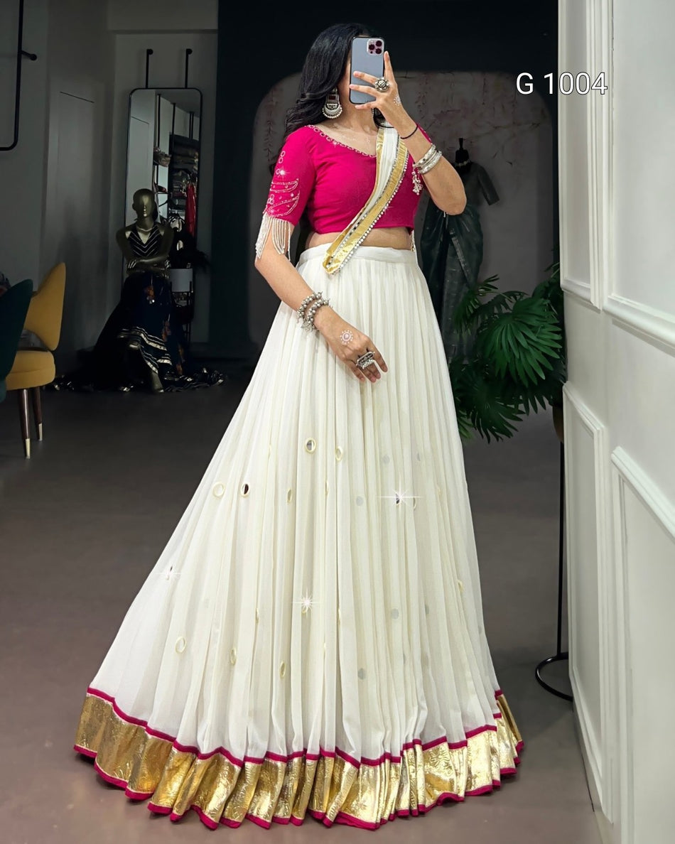 A woman takes a mirror selfie wearing a traditional Indian outfit consisting of a white flared skirt with gold and pink borders, a bright pink blouse with gold embroidery, and a gold sash. The setting appears to be an indoor boutique or dressing room with a mirror, chair, and plant in the background.