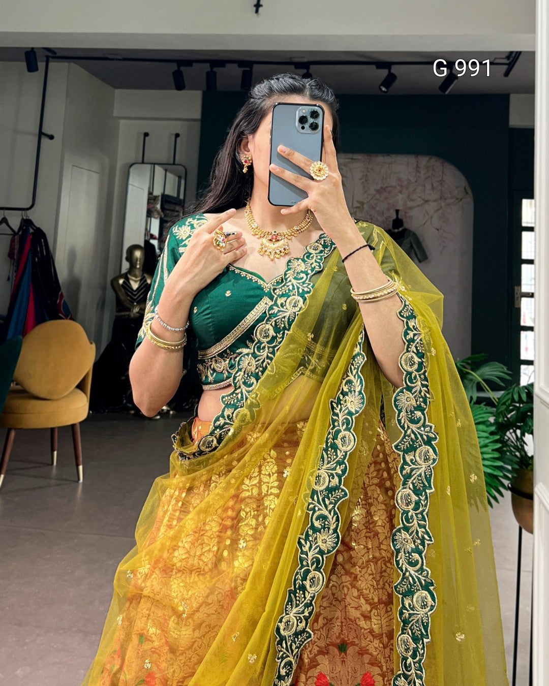 A woman takes a mirror selfie wearing a traditional Indian lehenga choli set. The outfit features a deep green embroidered choli with a yellow and gold embroidered lehenga and a sheer yellow dupatta with green floral borders.