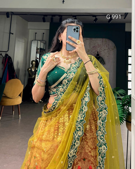 A woman takes a mirror selfie wearing a traditional Indian lehenga choli set. The outfit features a deep green embroidered choli with a yellow and gold embroidered lehenga and a sheer yellow dupatta with green floral borders.