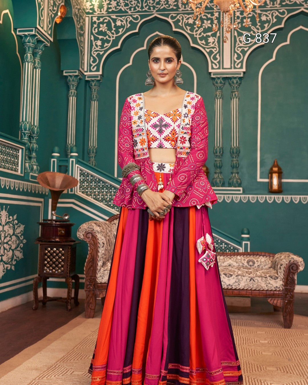 Woman in a colorful traditional outfit standing in a decorated room with ornate walls and furniture.