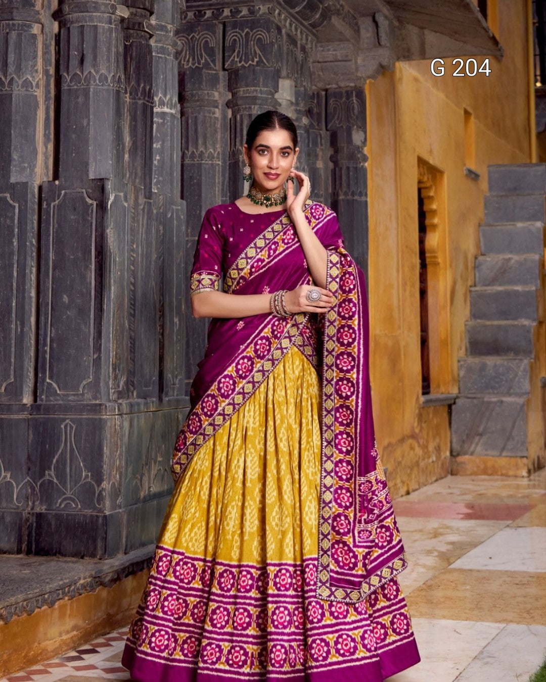 Woman in a traditional purple and yellow saree standing in front of an architectural background.
