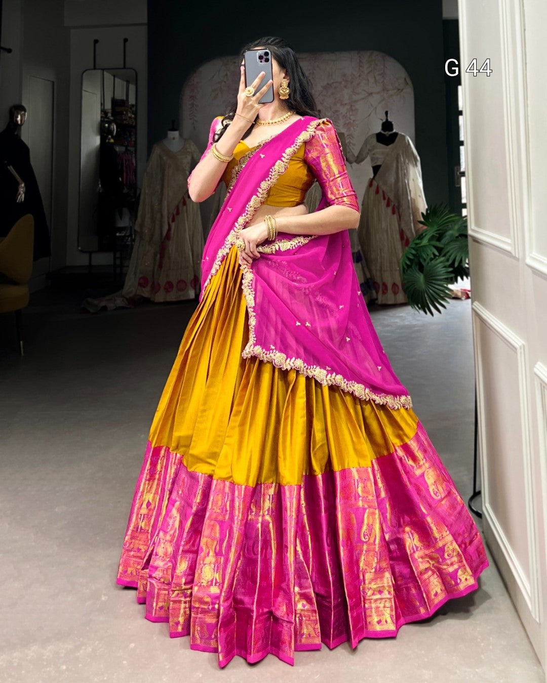 Woman wearing a yellow and pink saree taking a mirror selfie in a room.