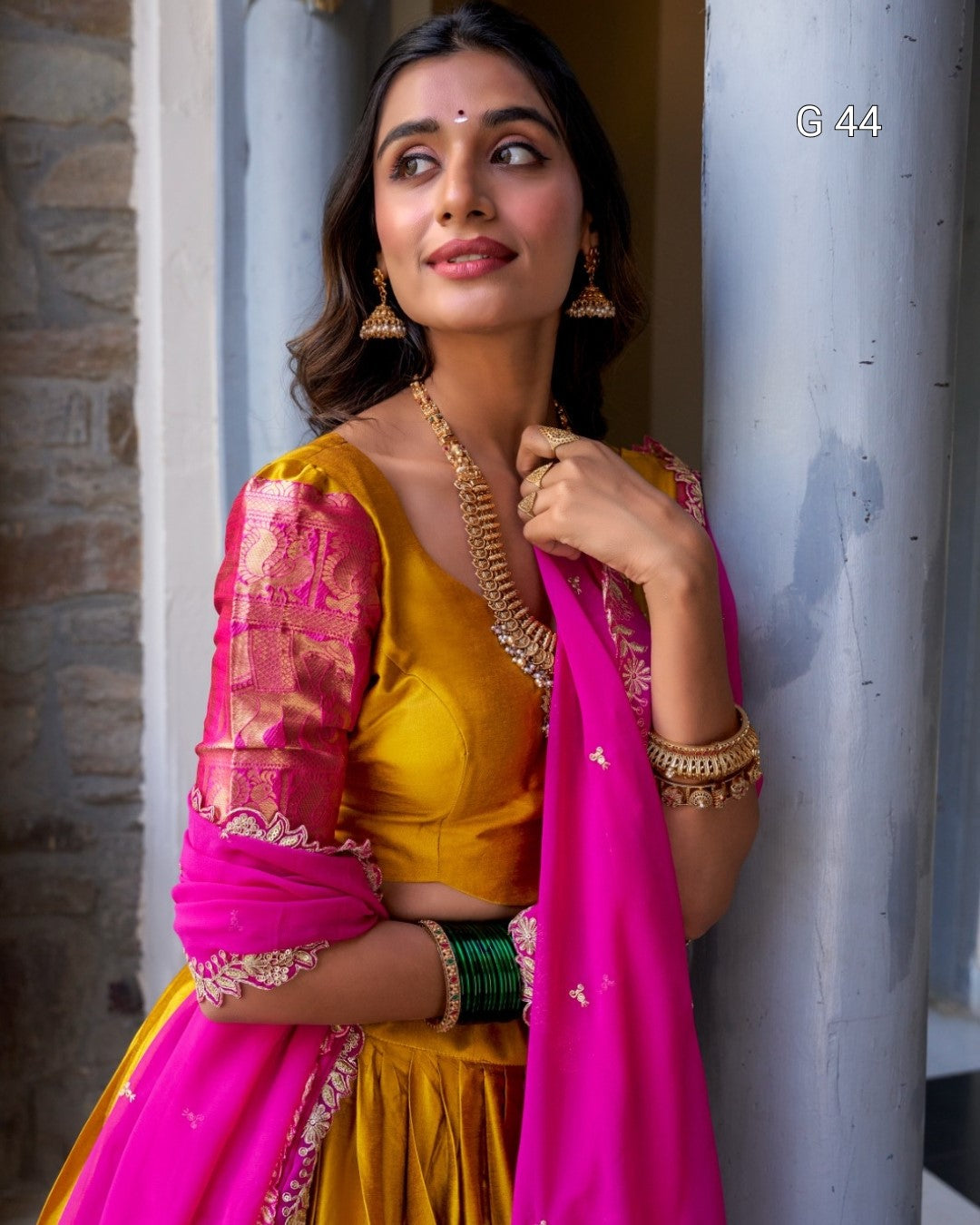 Woman in a yellow and pink traditional outfit with jewelry, leaning against a wall.
