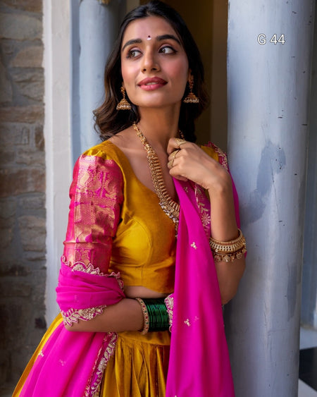 Woman in a yellow and pink traditional outfit with jewelry, leaning against a wall.