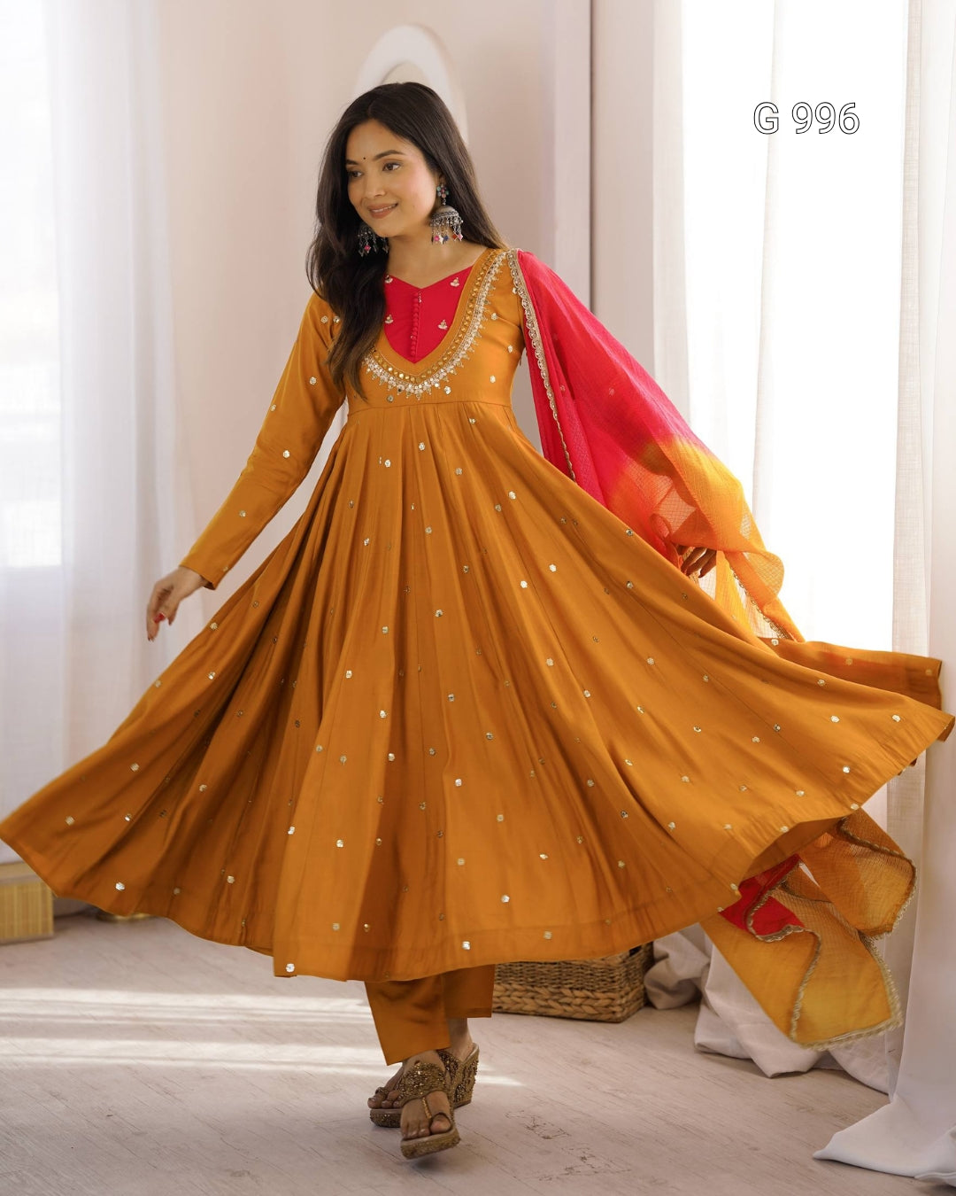 A woman modeling a traditional Indian outfit featuring a mustard yellow anarkali dress with silver mirror work, a red embroidered neckline, and a red and yellow gradient dupatta, set against a bright indoor background with white curtains.