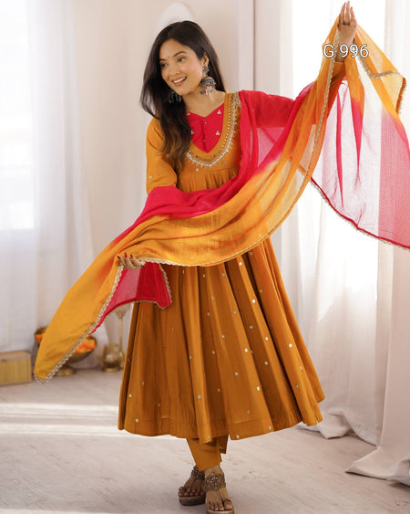 A woman modeling a traditional Indian outfit consisting of a mustard yellow Anarkali kurta and matching pants, with a bright red and yellow gradient dupatta draped over her shoulders. She is standing in a brightly lit room with white curtains.