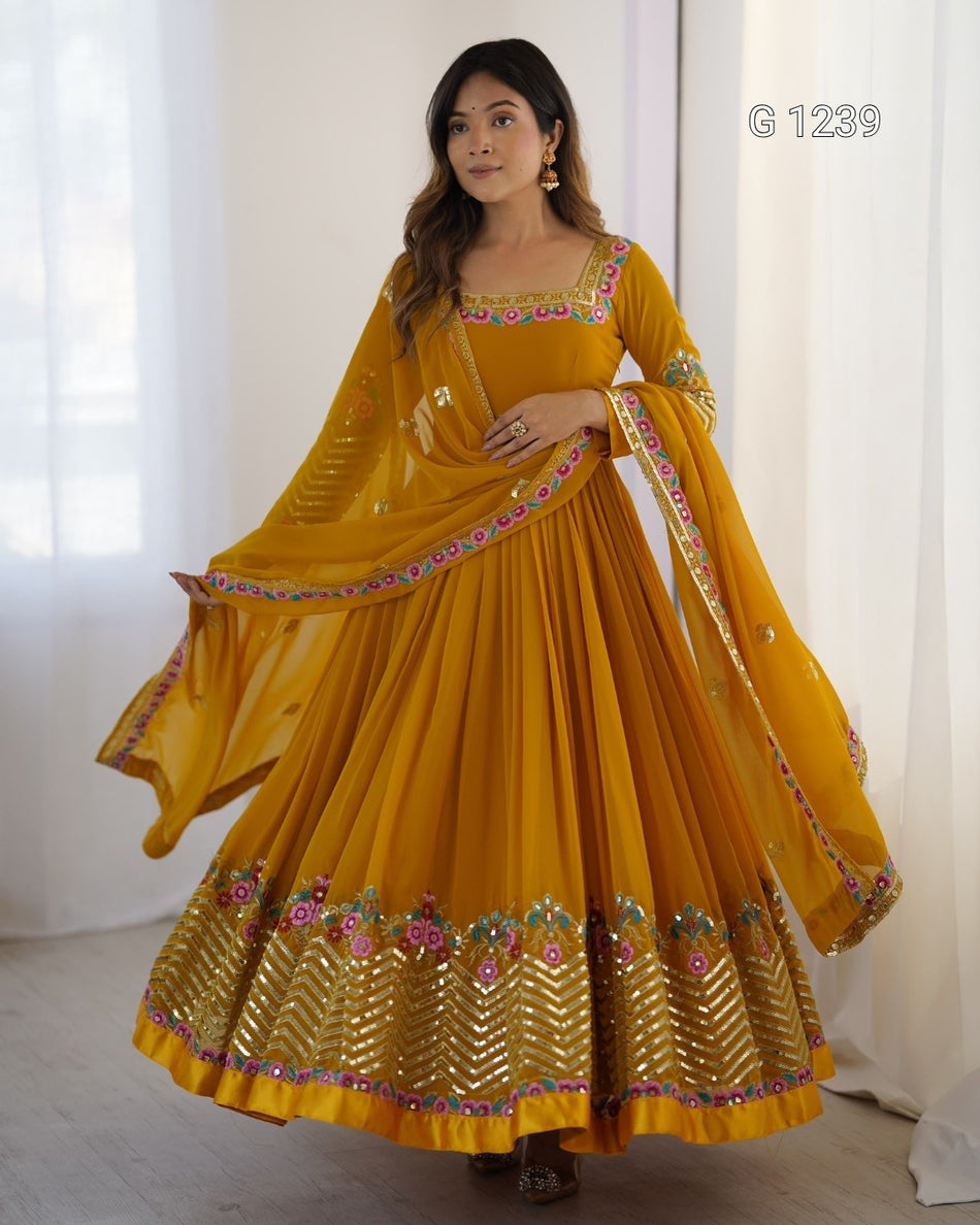 A woman modeling a traditional Indian outfit consisting of a mustard yellow anarkali skirt with gold embroidery, a matching blouse, and a dupatta, set against a soft white curtain background.