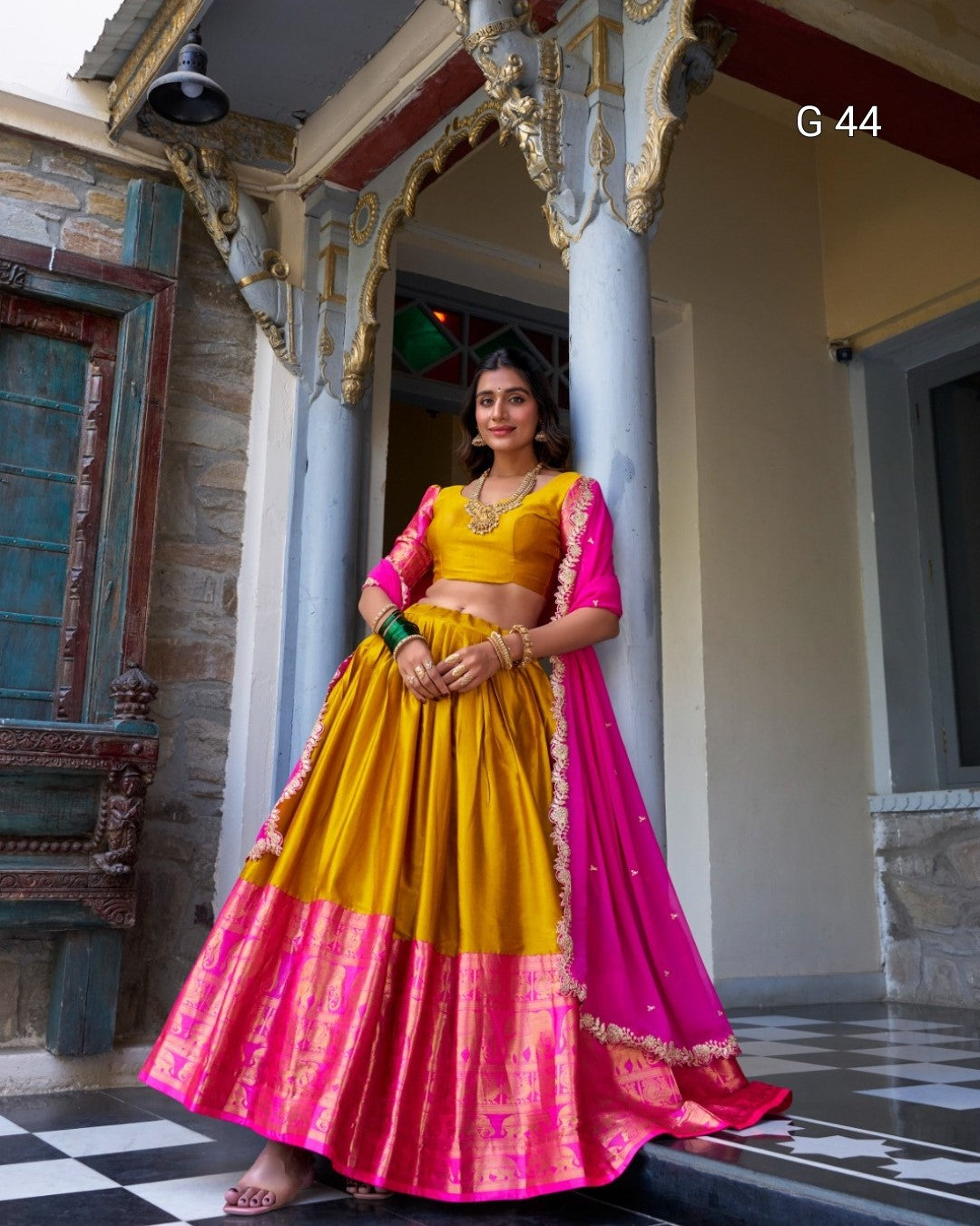 Woman in a yellow and pink traditional outfit standing in an ornate indoor setting.