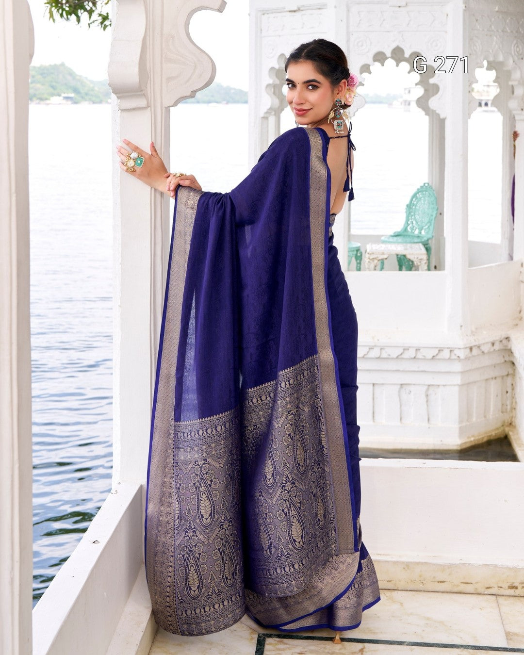 A woman models a royal blue saree with a wide beige border and a heavily embroidered beige and silver border, standing on a white architectural balcony overlooking water.