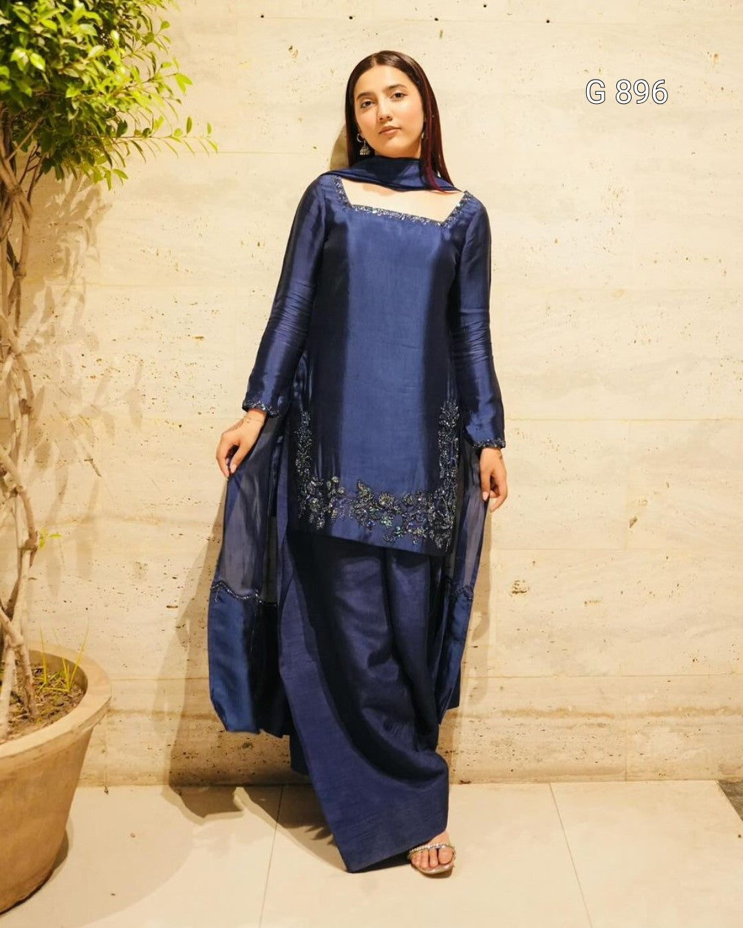 Woman wearing a blue traditional outfit with intricate designs, standing against a textured wall.