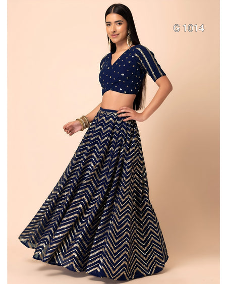 A woman modeling a navy blue traditional outfit with a matching flared skirt featuring a gold chevron pattern and a short-sleeve crop top with gold embellishments, set against a solid beige background.
