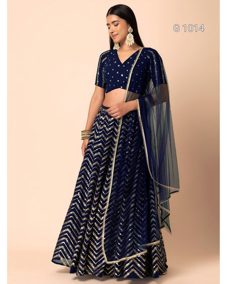 A woman modeling a navy blue traditional lehenga choli set with silver zari embroidery and a sheer dupatta, posed against a solid beige studio background.