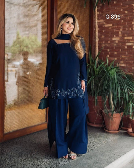 Woman in a navy blue outfit standing outdoors with plants and a textured wall in the background.