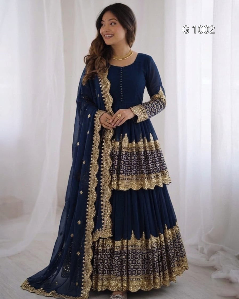 A woman modeling a navy blue traditional Anarkali gown with intricate gold embroidery and a matching dupatta, set against a soft white curtain backdrop.