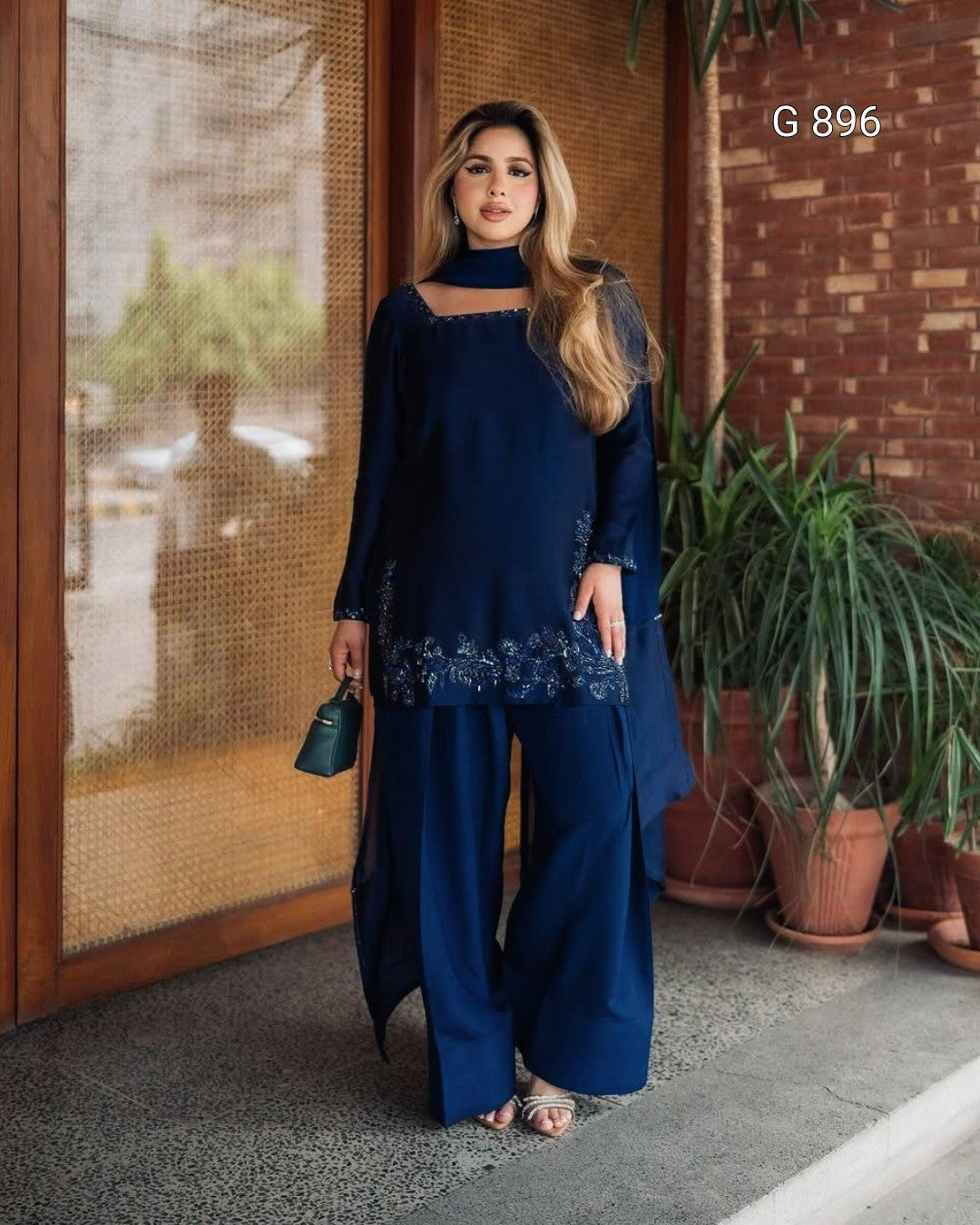 Woman in a blue outfit standing outdoors with plants and a brick wall in the background