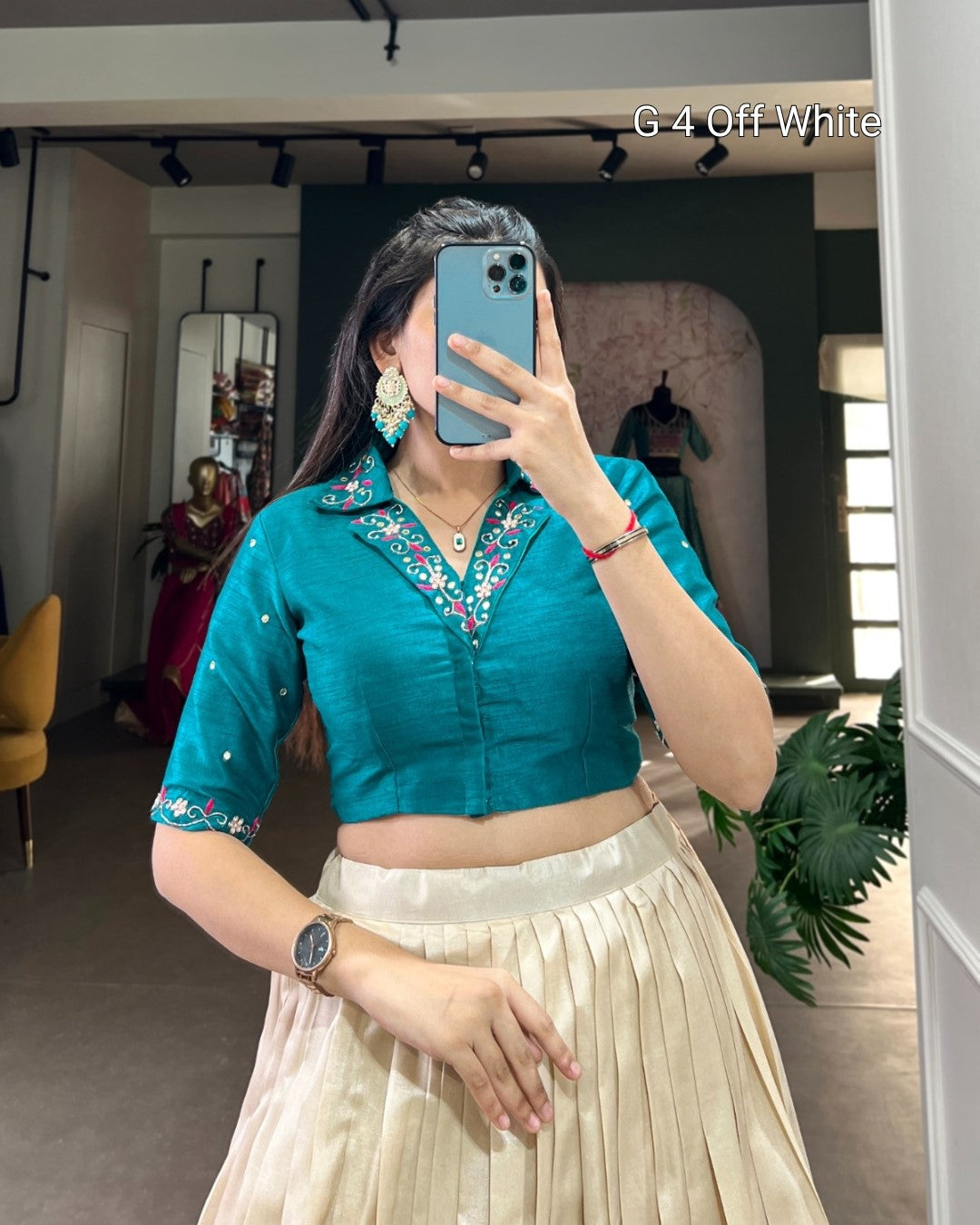 Woman taking a mirror selfie wearing a teal blouse with floral embroidery and a beige skirt.