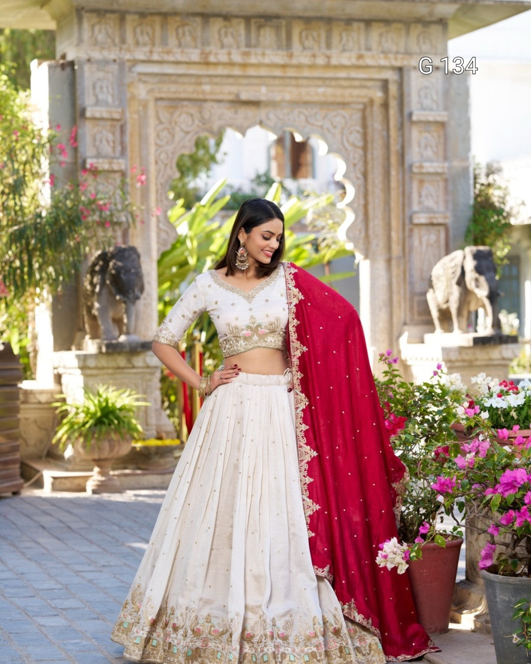 Woman in a white and red traditional outfit standing in a garden with statues and plants.