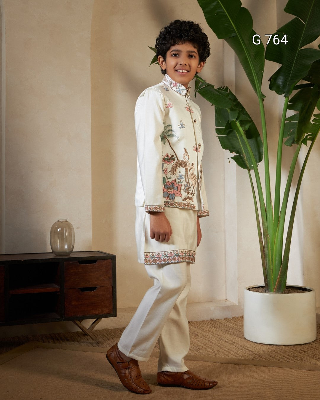 Young boy in traditional outfit standing in a room with a plant and wooden cabinet.