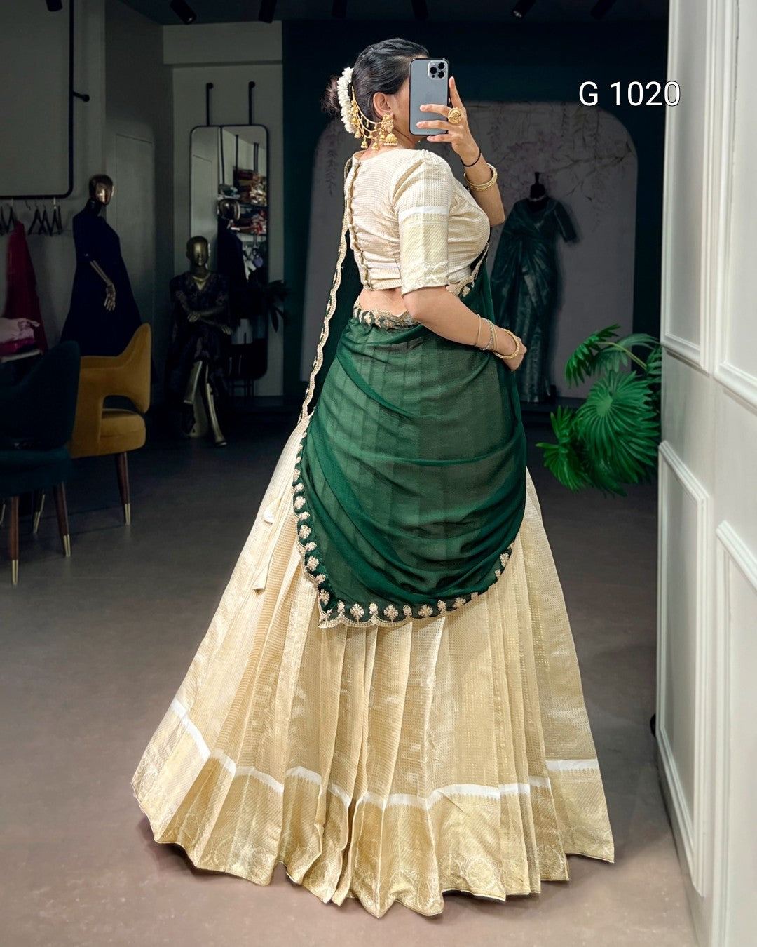 A woman wearing a traditional Indian lehenga choli dress, photographed in a mirror selfie inside what appears to be a boutique or showroom. The outfit features a cream-colored flared skirt (lehenga) with white border details and a dark green draped blouse and dupatta (sari) with a decorative border of small white flowers.
